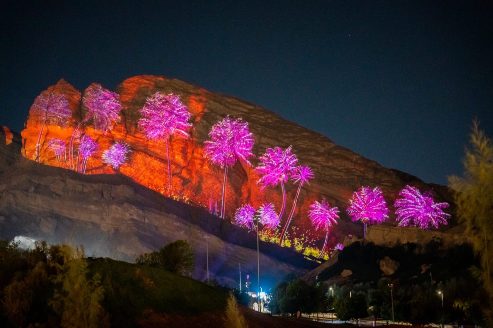 A rocky mountainside in Al Ain lit in pink and orange by a large-scale projection at night, with trees in the foreground