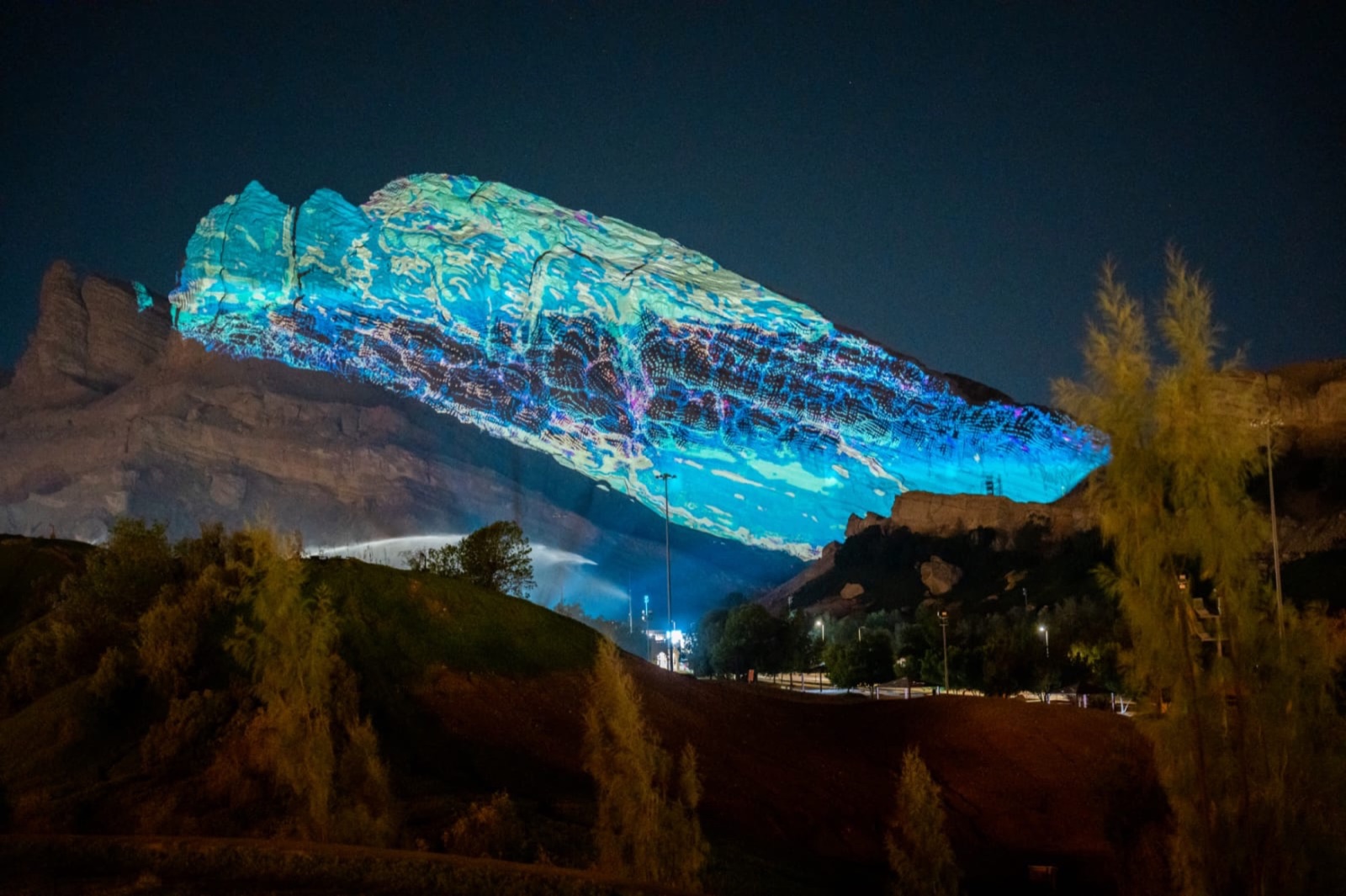 A rocky mountainside in Al Ain lit in pink and orange by a large-scale projection at night, with trees in the foreground