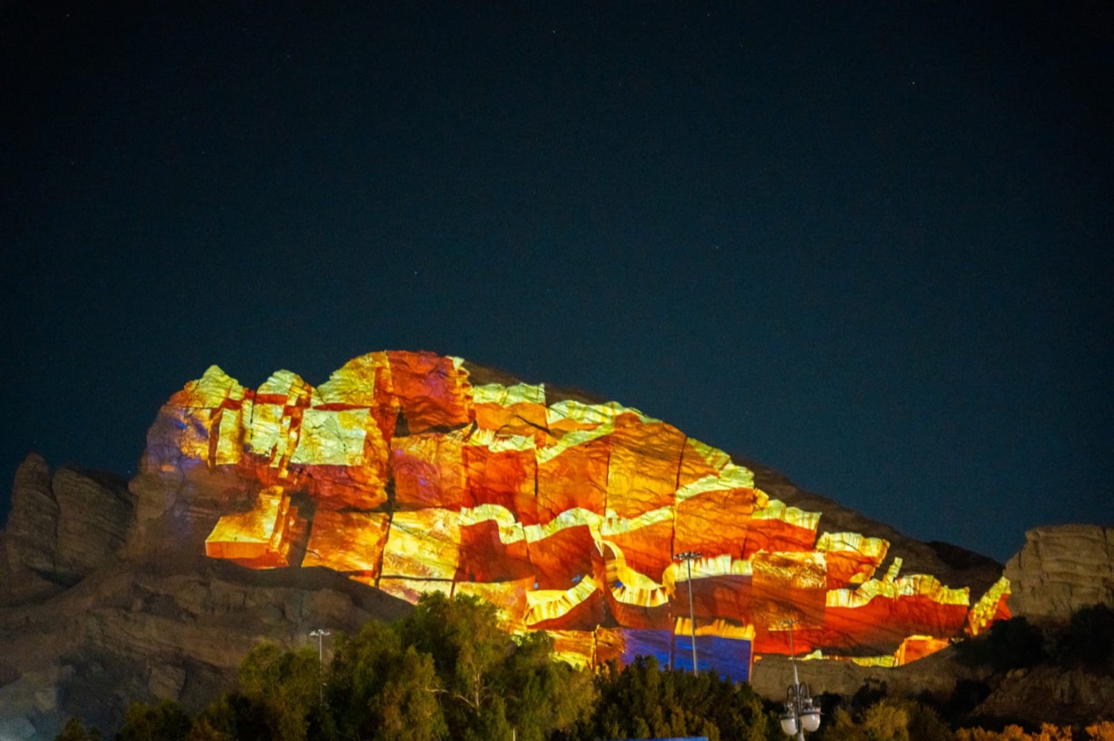 A rocky mountainside in Al Ain lit in pink and orange by a large-scale projection at night, with trees in the foreground