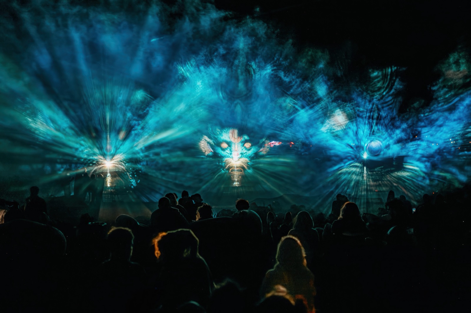 A crowd at night in front of three fan-shaped screens of water and fog on which giant blue faces of light appear — BEYOND by RE:SORB at Boom Festival