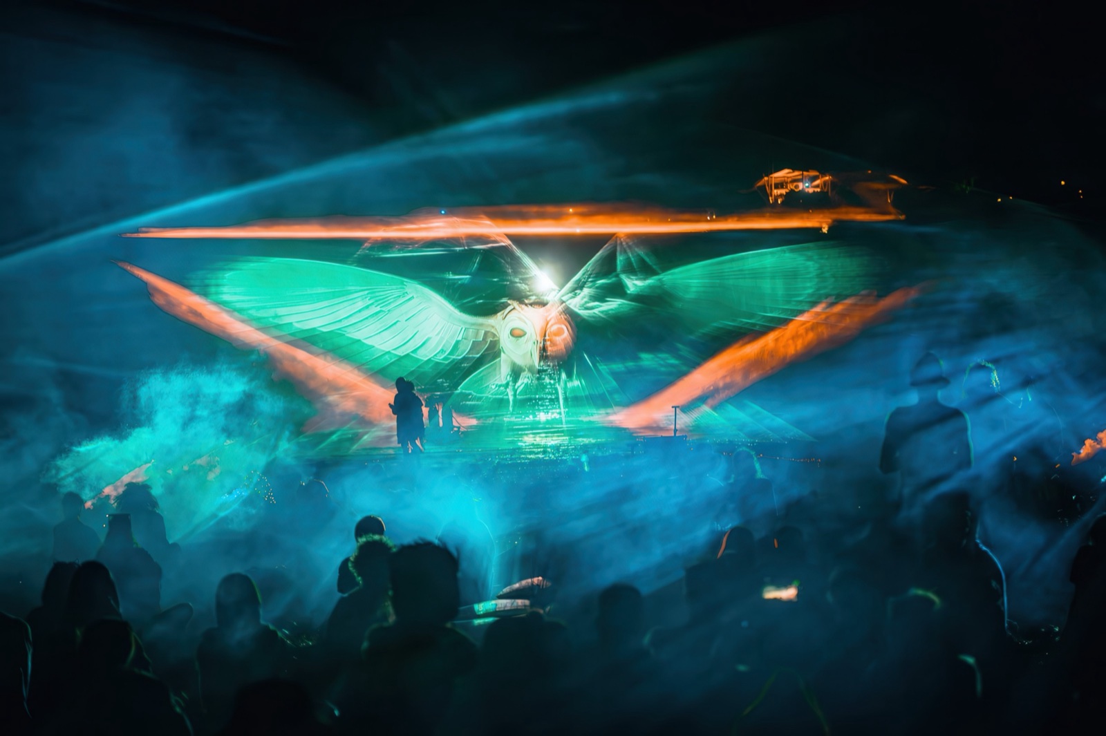 A crowd at night in front of three fan-shaped screens of water and fog on which giant blue faces of light appear — BEYOND by RE:SORB at Boom Festival