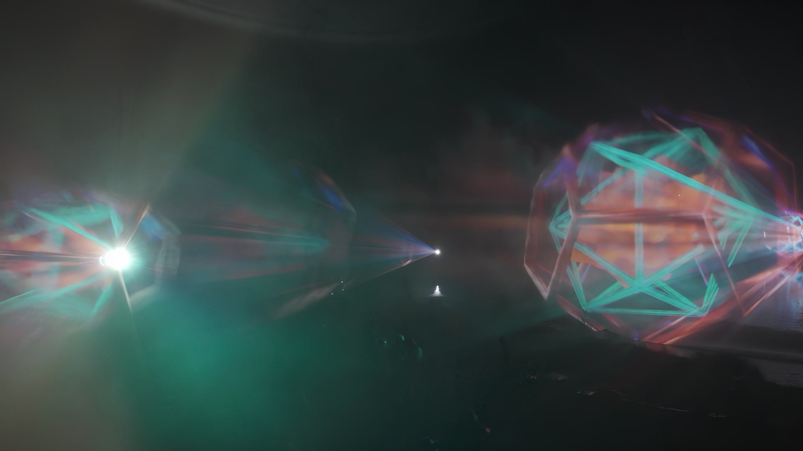 Beams of blue and orange light passing through layered water and mist screens at night, with festival visitors silhouetted in front of the Depth/100 installation at Ozora