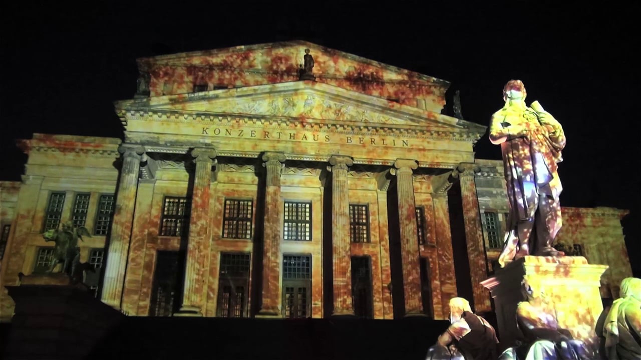 The columned façade of the Konzerthaus at Gendarmenmarkt in Berlin lit by a colourful projection mapping with the Schiller monument in front