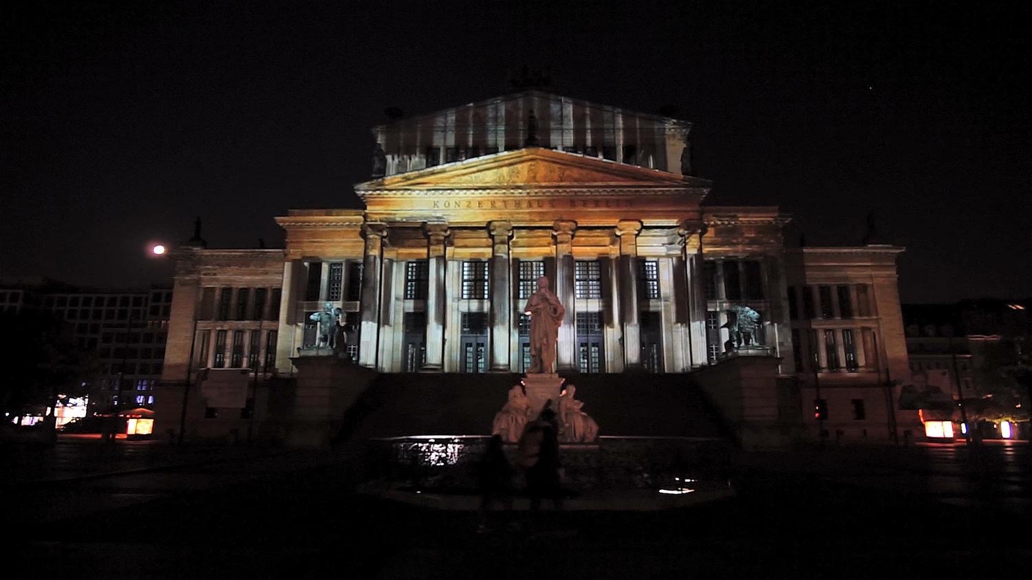 The columned façade of the Konzerthaus at Gendarmenmarkt in Berlin lit by a colourful projection mapping with the Schiller monument in front