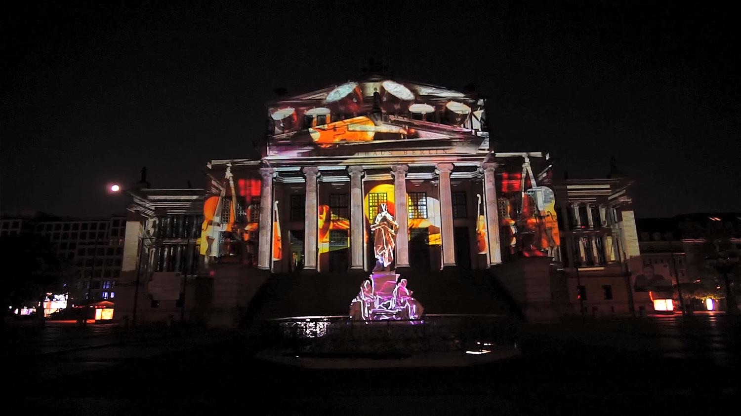 The columned façade of the Konzerthaus at Gendarmenmarkt in Berlin lit by a colourful projection mapping with the Schiller monument in front