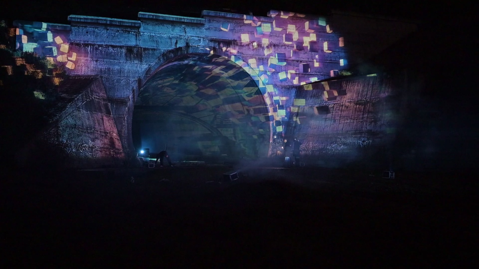 A stone railway tunnel portal in Tbilisi covered in a projection mapping of coloured tiles, with a hologram of light appearing in the tunnel mouth
