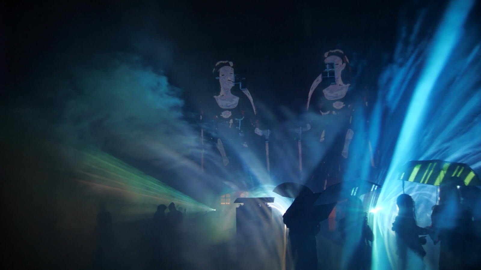 A crowd at night in front of a mapped building at Boom Festival, with beams of blue and white light passing through fog in front of the façade