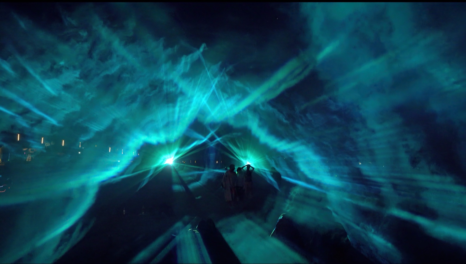 A crowd at night in front of a mapped building at Boom Festival, with beams of blue and white light passing through fog in front of the façade