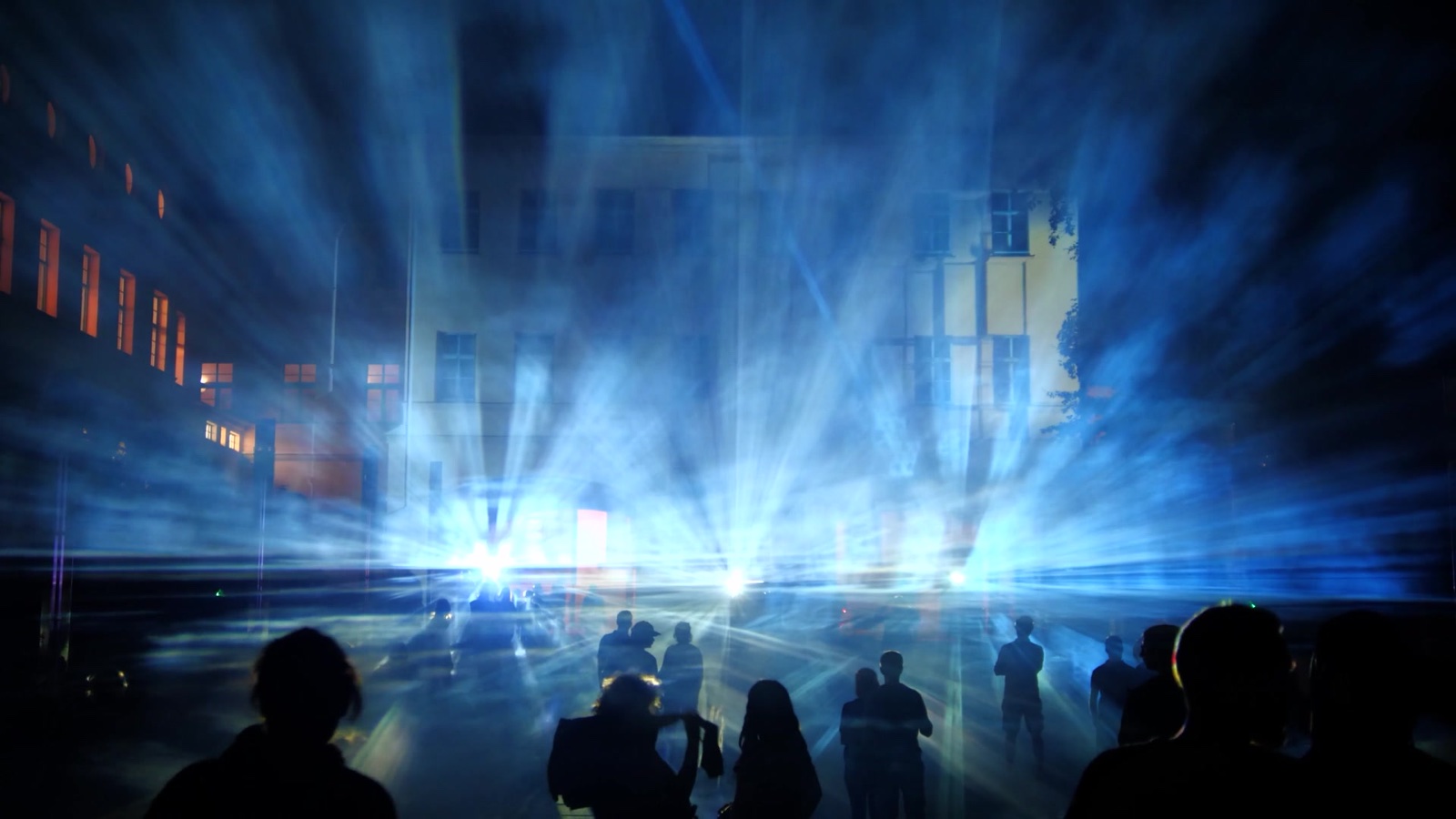 A crowd at night in front of a mapped building at Boom Festival, with beams of blue and white light passing through fog in front of the façade
