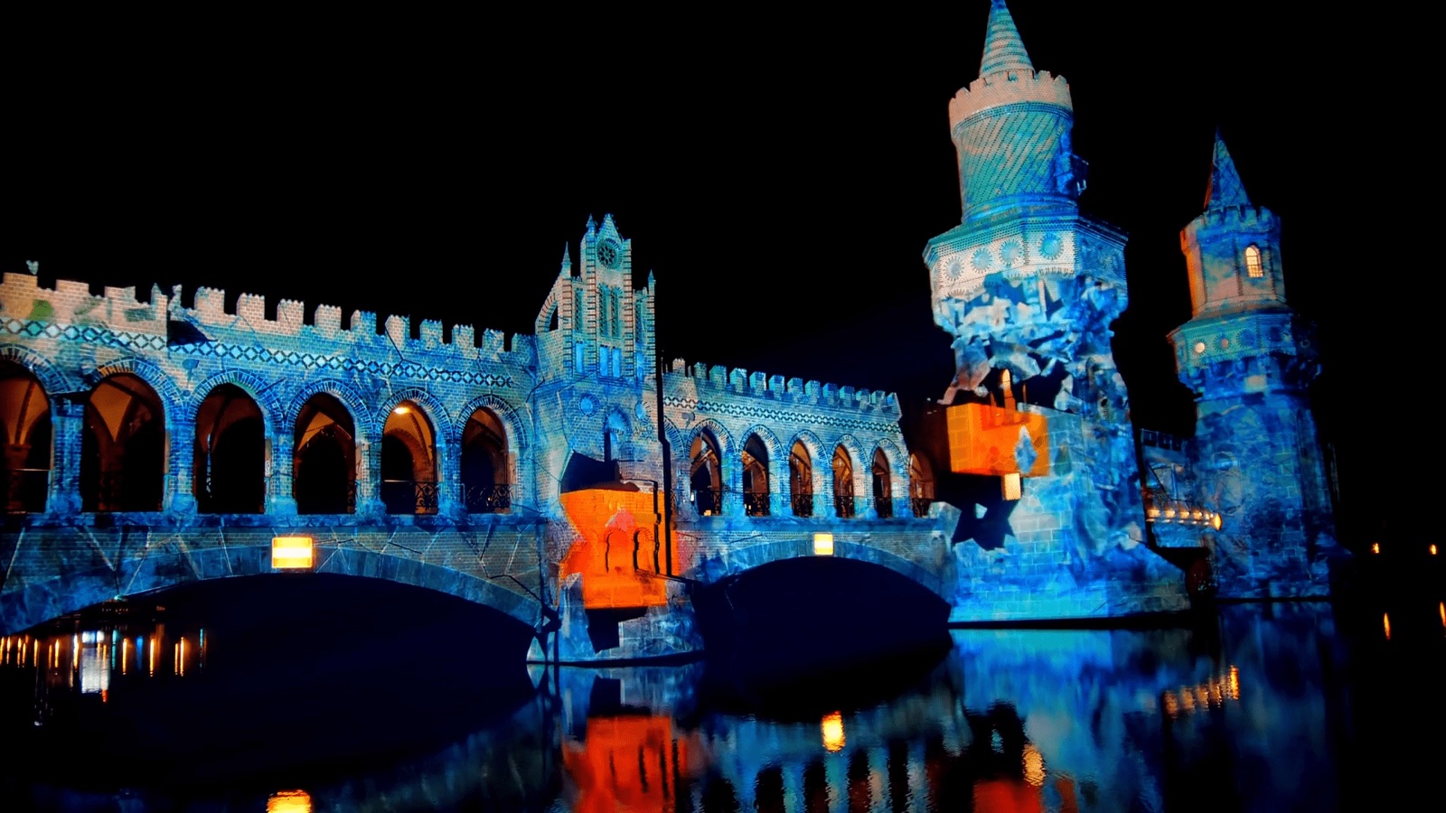 The twin brick towers of the Oberbaumbrücke in Berlin illuminated in orange, red and blue by a projection mapping, reflected in the river Spree
