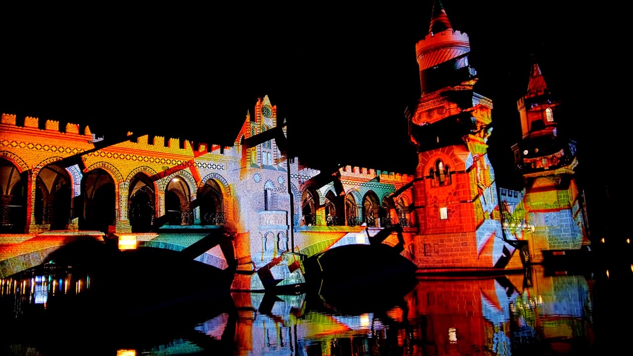 The twin brick towers of the Oberbaumbrücke in Berlin illuminated in orange, red and blue by a projection mapping, reflected in the river Spree