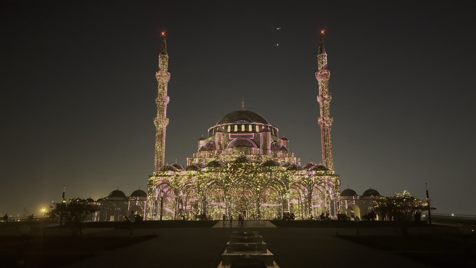 A domed mosque with two minarets in Sharjah covered in a fine golden projection mapping at night