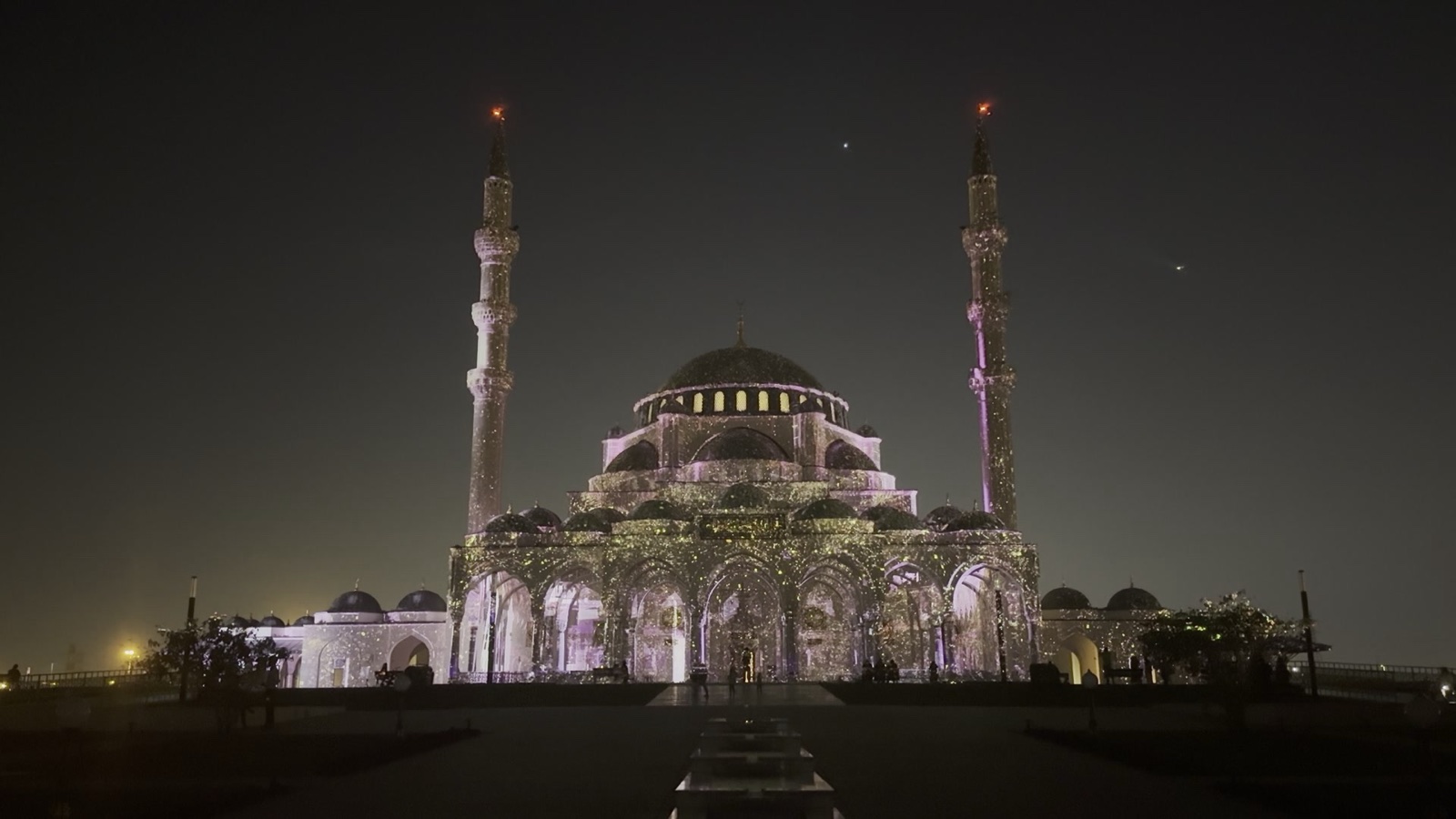 A domed mosque with two minarets in Sharjah covered in a fine golden projection mapping at night