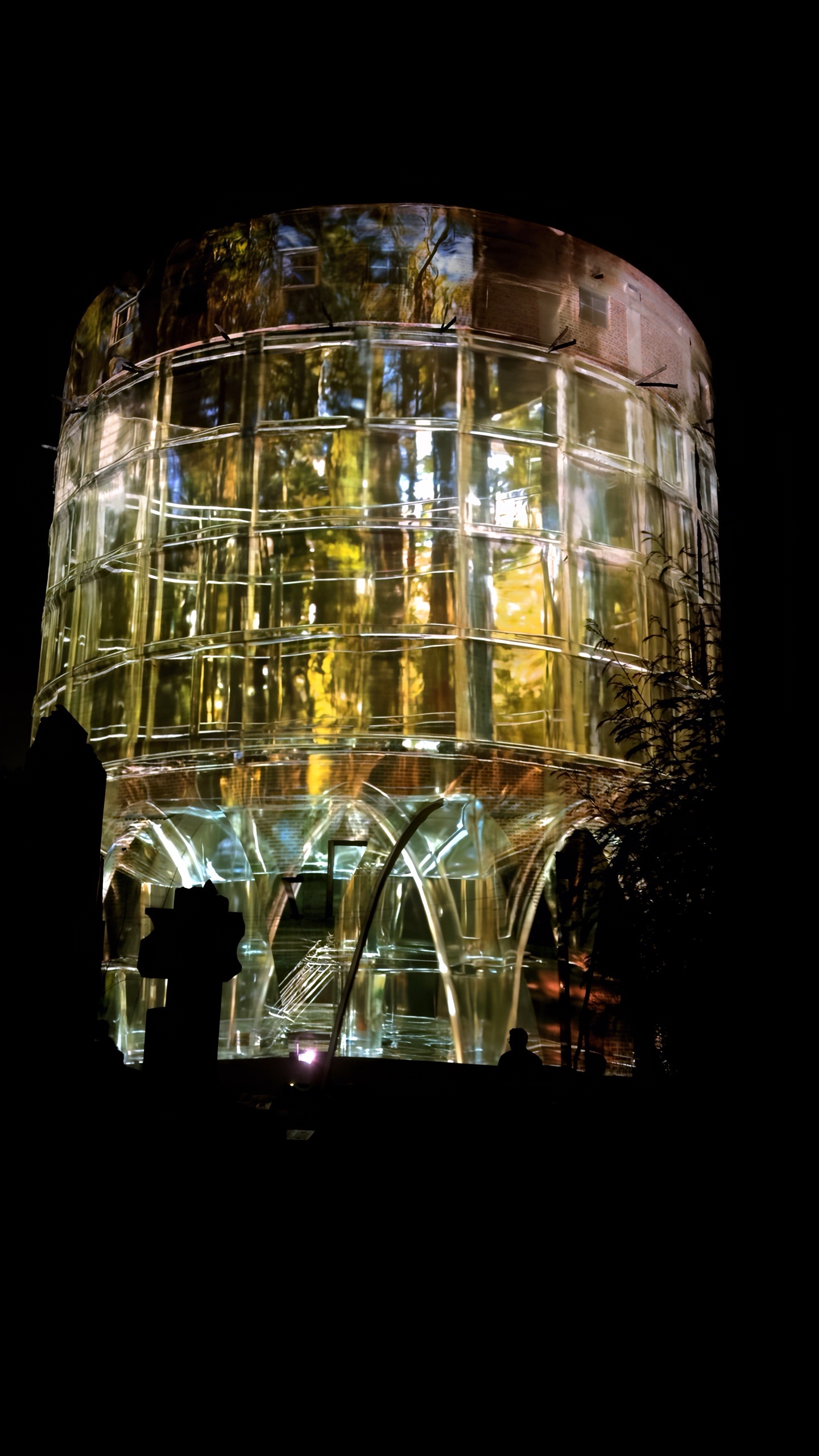 The 24-metre concrete silo of the Alembic Glass Factory in Bengaluru rendered as a transparent lattice by projection, with an amber interior glowing inside and holographic fog around it